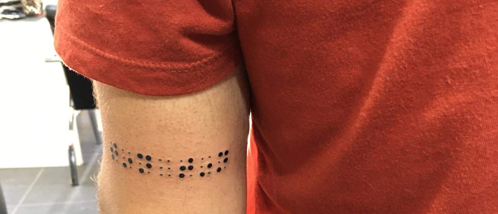 Yakın plan çekimde Kol üzerindeki Braille ile dövme olarak yazılmış tın tın yazısı görülmekte.